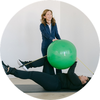 Woman being guided by instructor to use a green exercise ball with resistance bands in a gym setting.