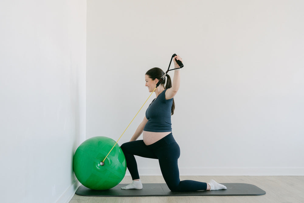 Pregnant woman exercising with a resistance band and exercise ball in a room with white walls
