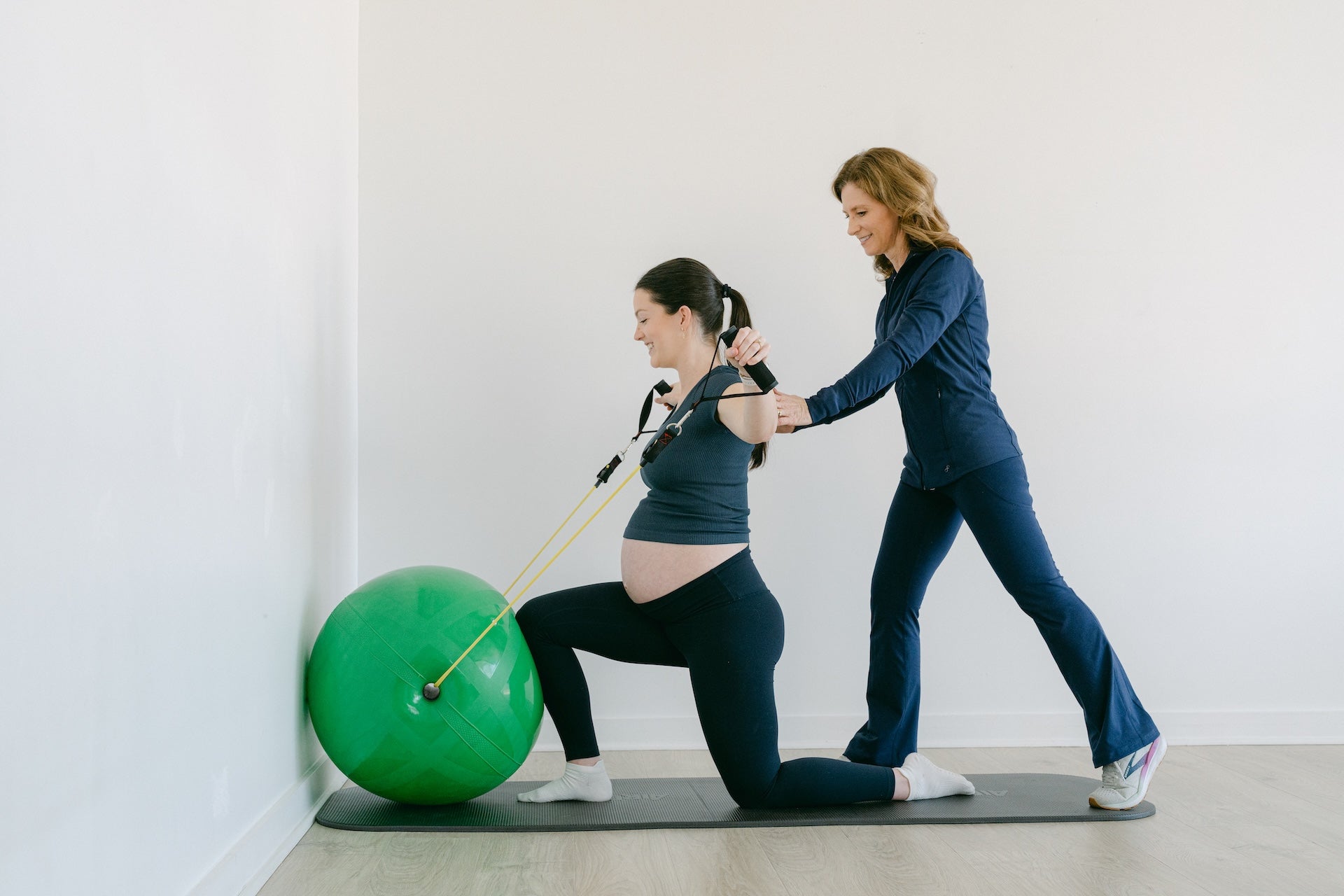 Pregnant woman exercising with a green ball and instructor assisting her on a white background