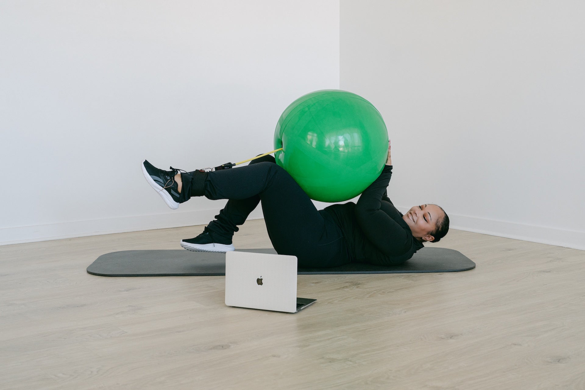Person exercising with a green fitness ball on a wooden floor, using a laptop.
