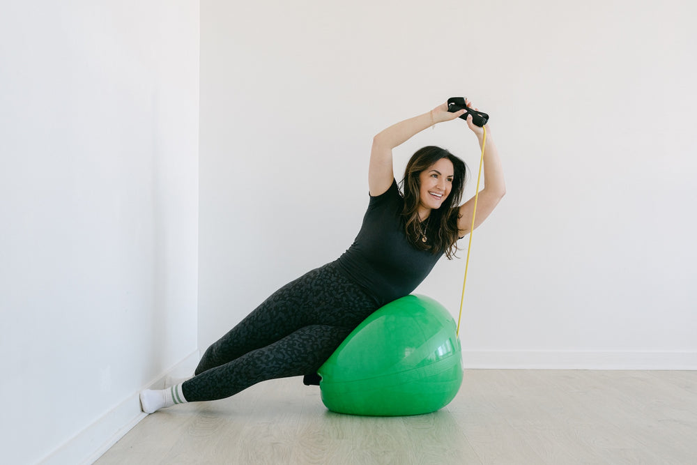 Woman exercising on a green ball with resistance bands in a room.