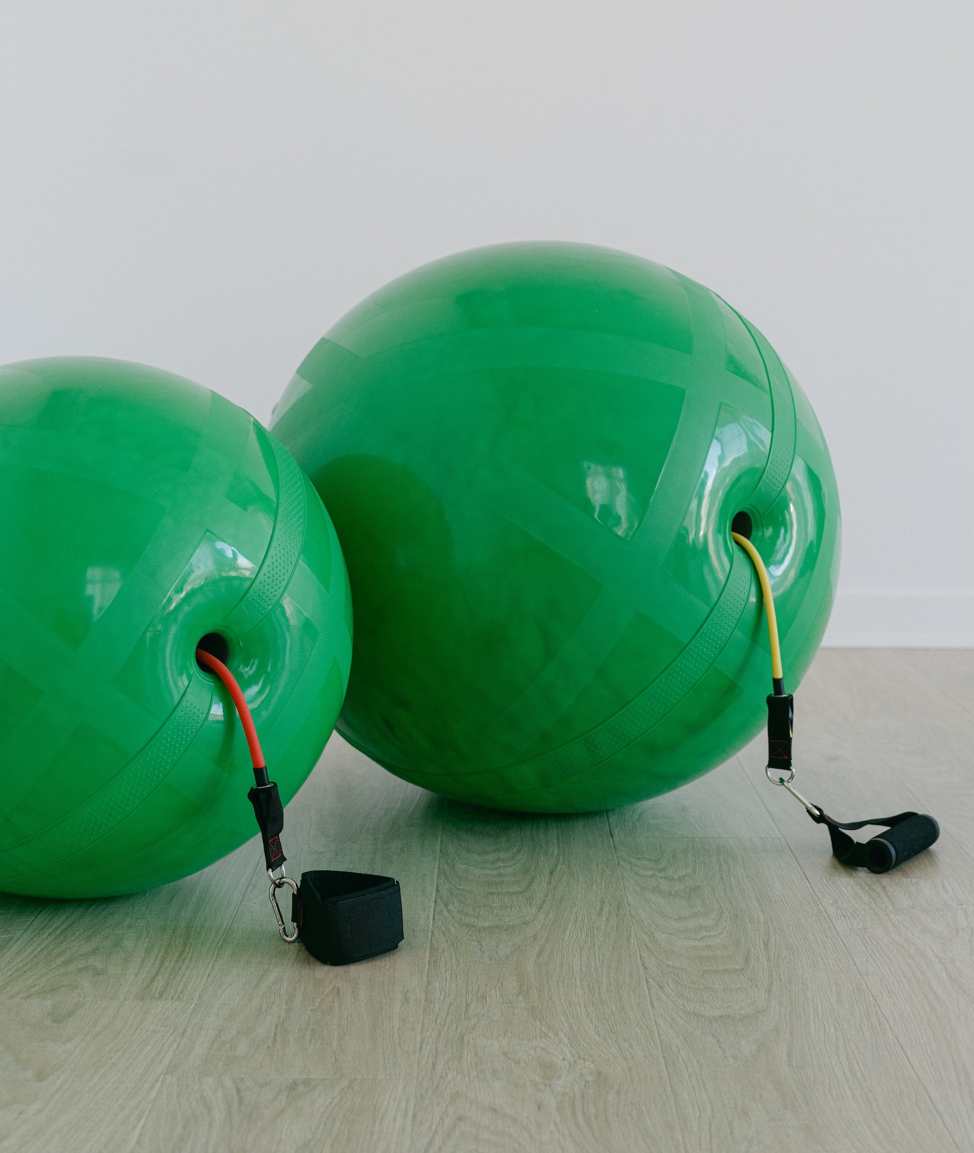 Two green exercise balls with resistance bands sitting next to eachother on a light wooden floor.
