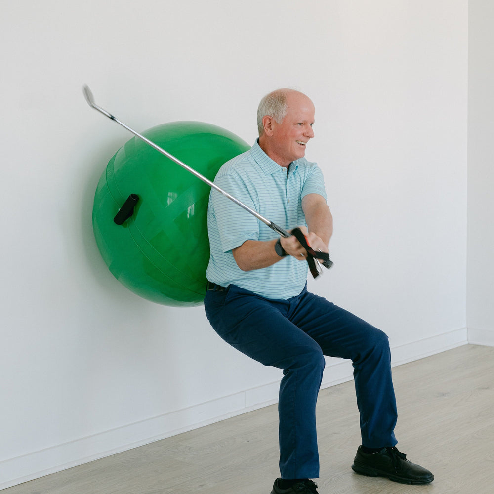 Man sitting on a large green ball with a pole attached, against a white wall.