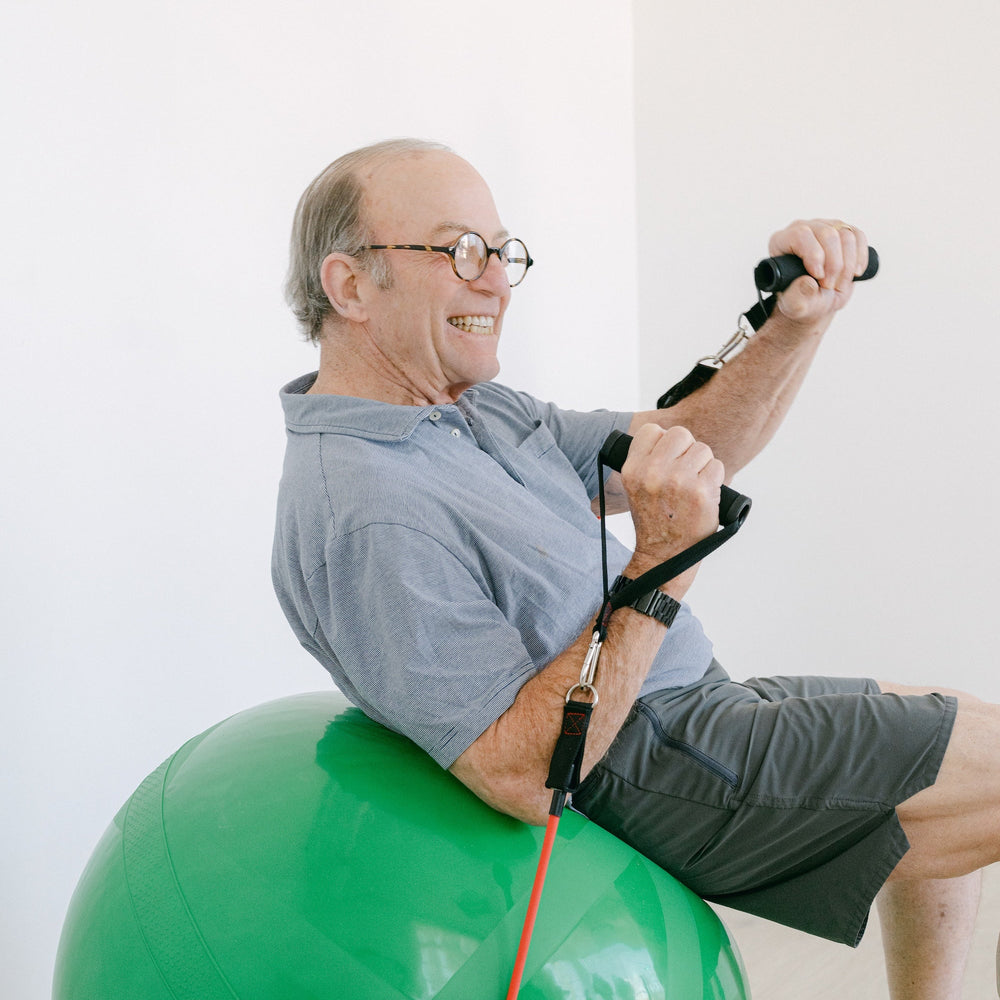 Man exercising on a green ball with resistance bands indoors.