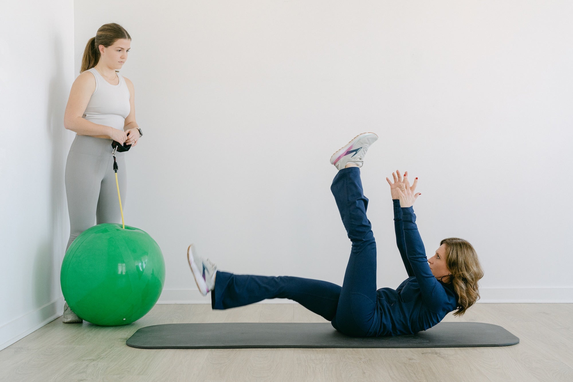 Woman exercising on a mat with a green exercise ball while another woman observes.